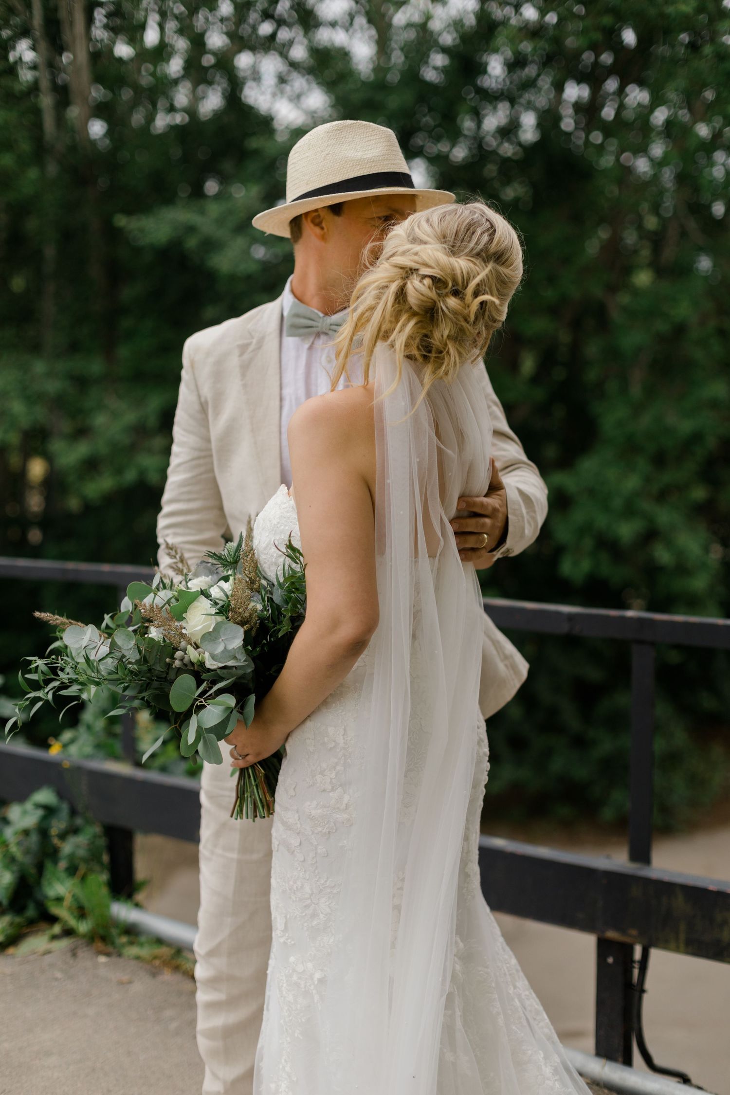 A couple in wedding attire embrace on a bridge, with the bride holding a lush greenery and white flower bouquet.