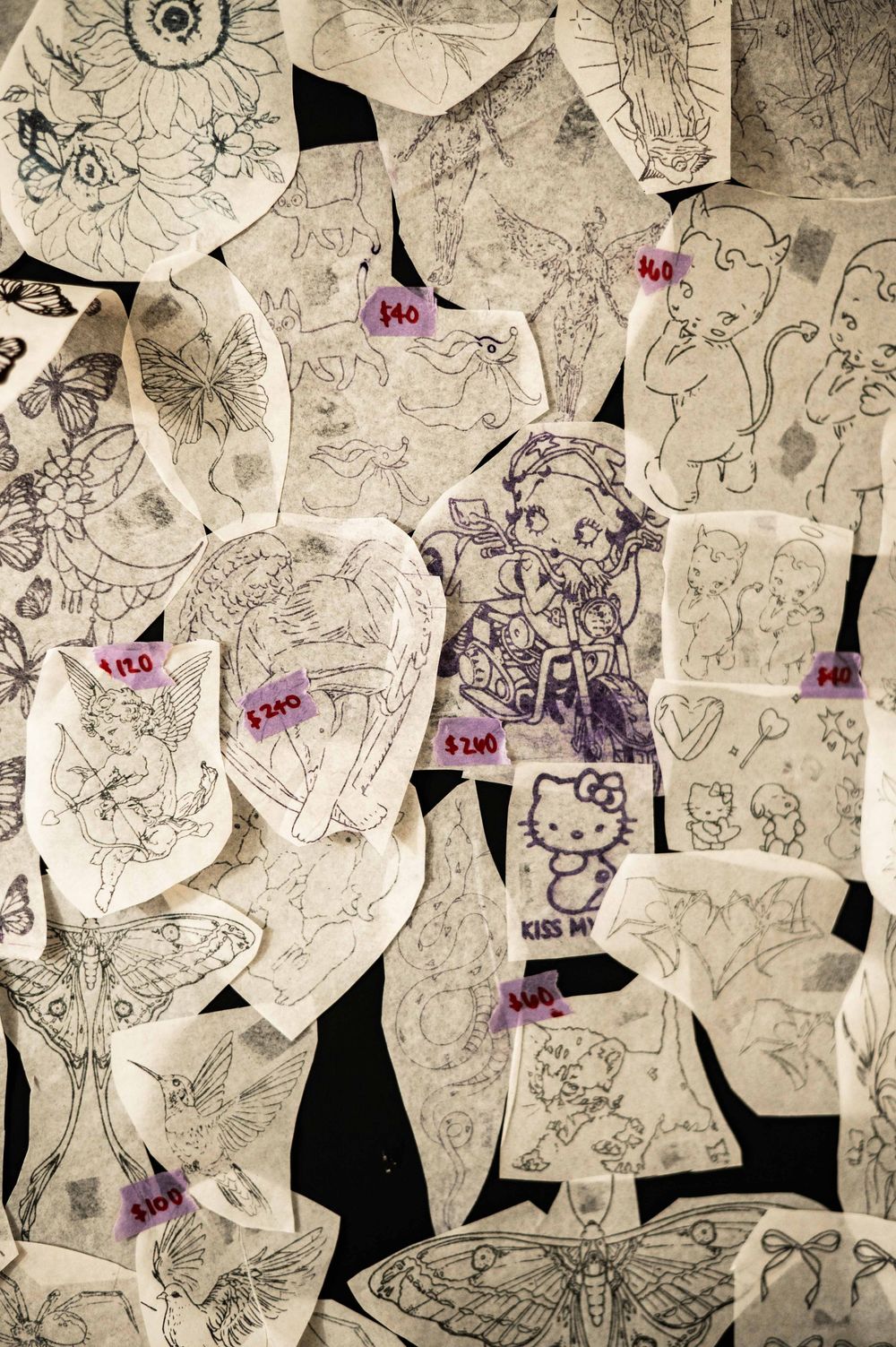 A wall covered with tattoo flash art designs including Hello Kitty, butterflies, and anime characters with price tags.