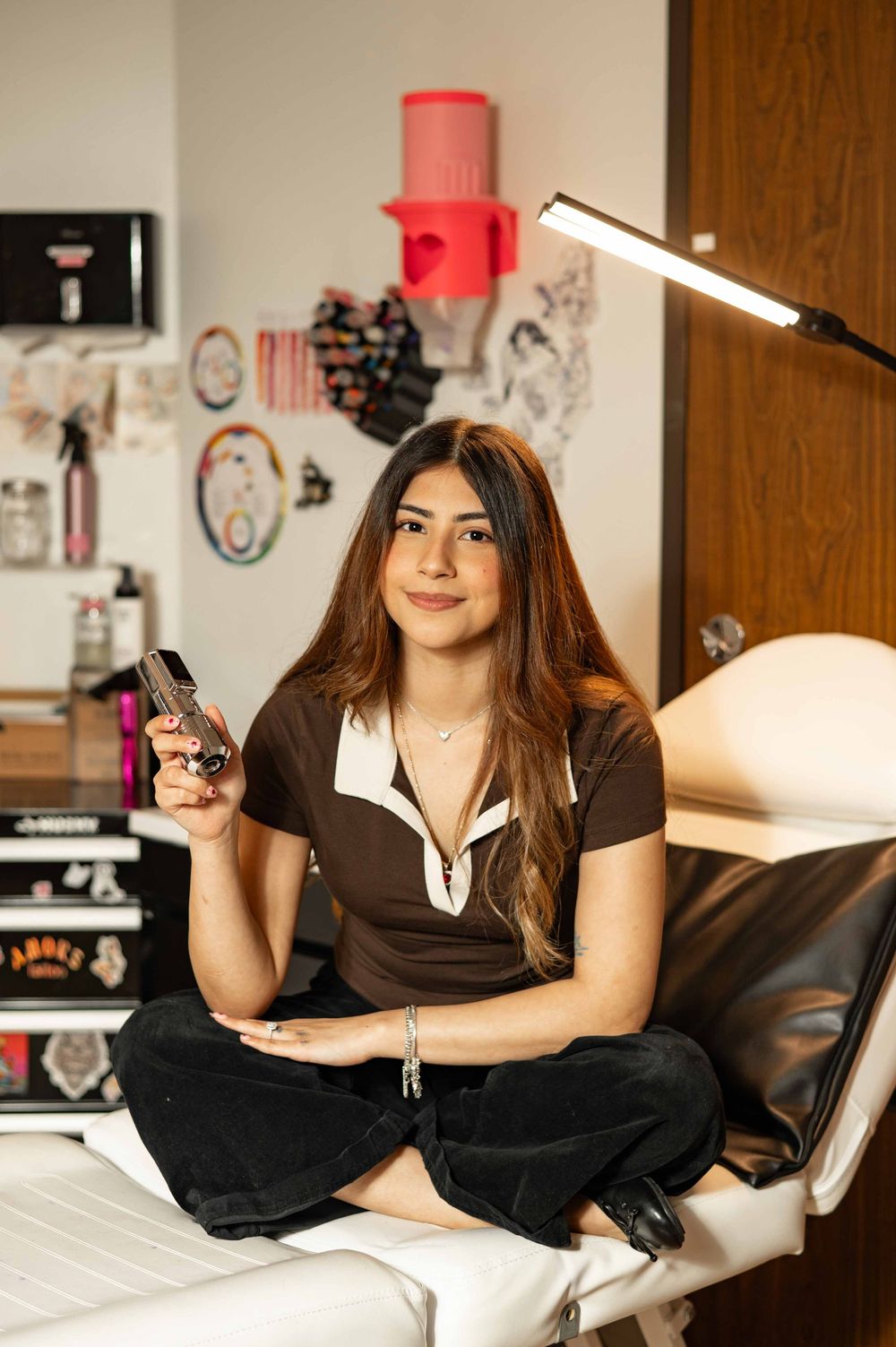 Young woman sitting cross-legged on a salon chair holding a phone in a tattoo studio setting.