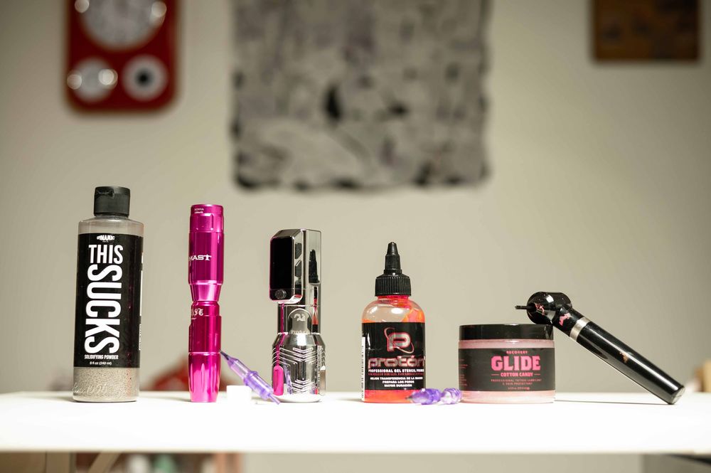 Assorted grooming products on a white shelf including This Sucks bottle, pink flashlight, hair clippers, oil, and styling glide.