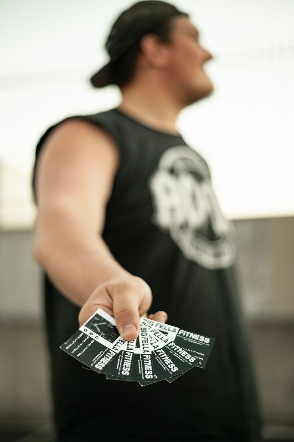 Muscular man in black sleeveless shirt holding out fitness business cards toward camera outdoors.