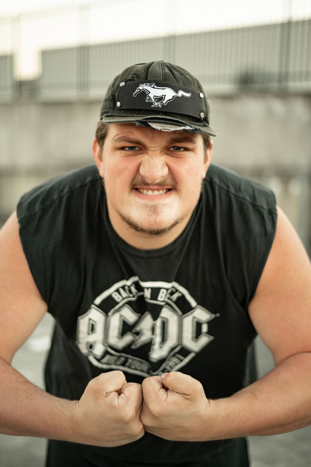 Young man in AC/DC shirt and Ford Mustang cap poses with clenched fists outdoors.