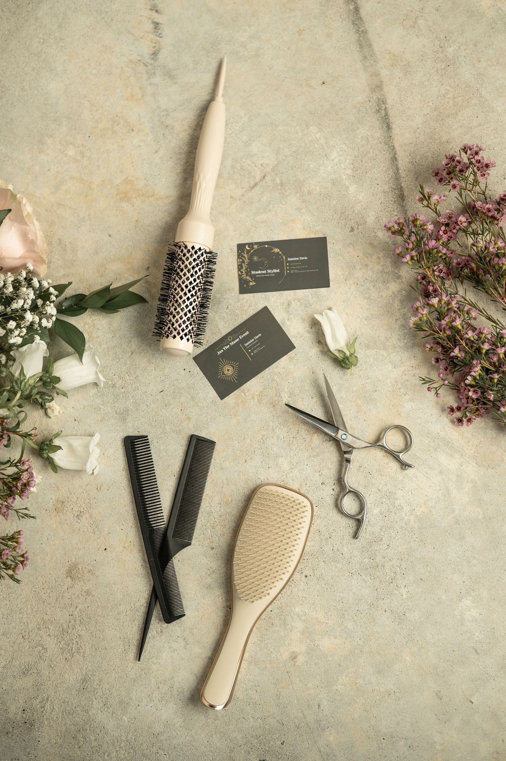 Professional hairstyling tools and pink flowers arranged with business cards on textured concrete background.