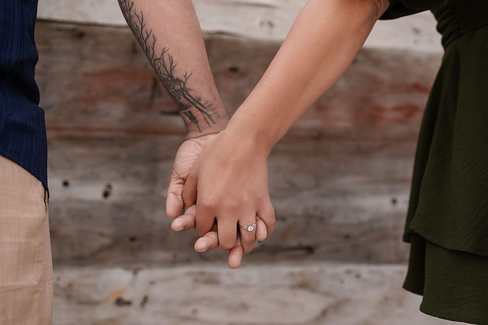 Two people hold hands and walk together in front of a rustic wooden fence.