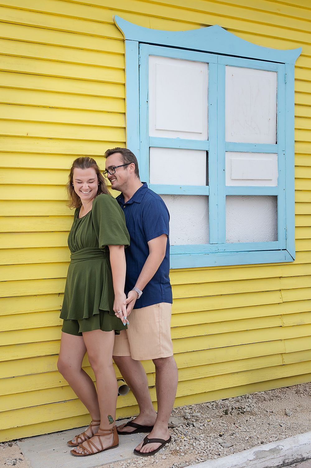 A couple poses playfully in front of a bright yellow building with blue trim windows.