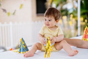 A toddler in a yellow shirt plays with colorful party decorations and streamers on a white table.
