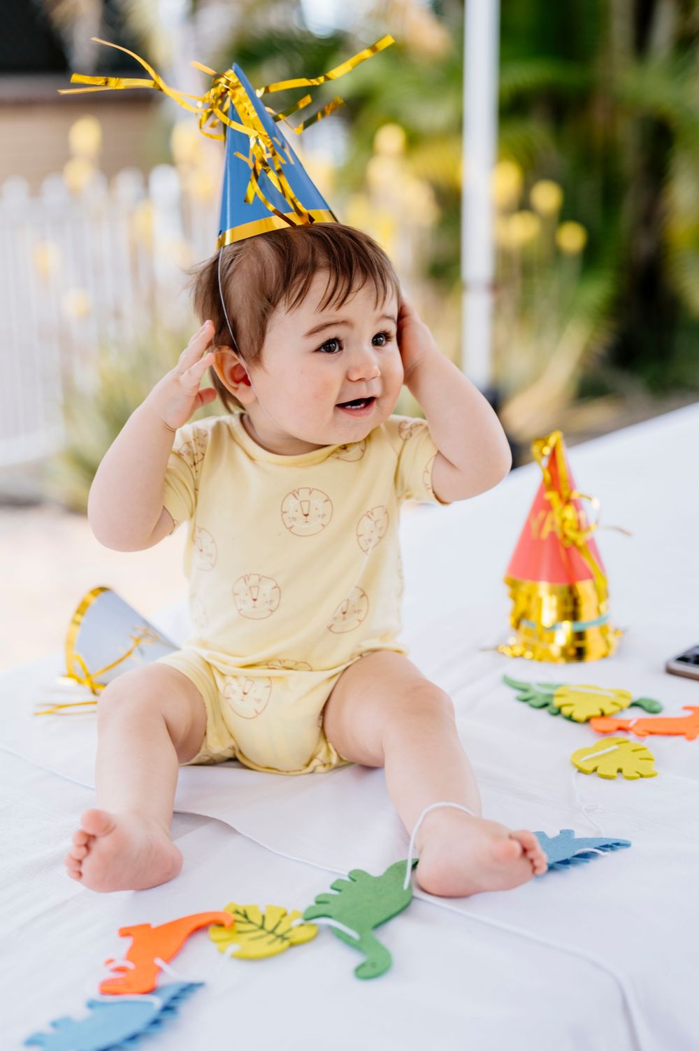 Baby wearing yellow romper and party hat sits on white surface with colorful party decorations.