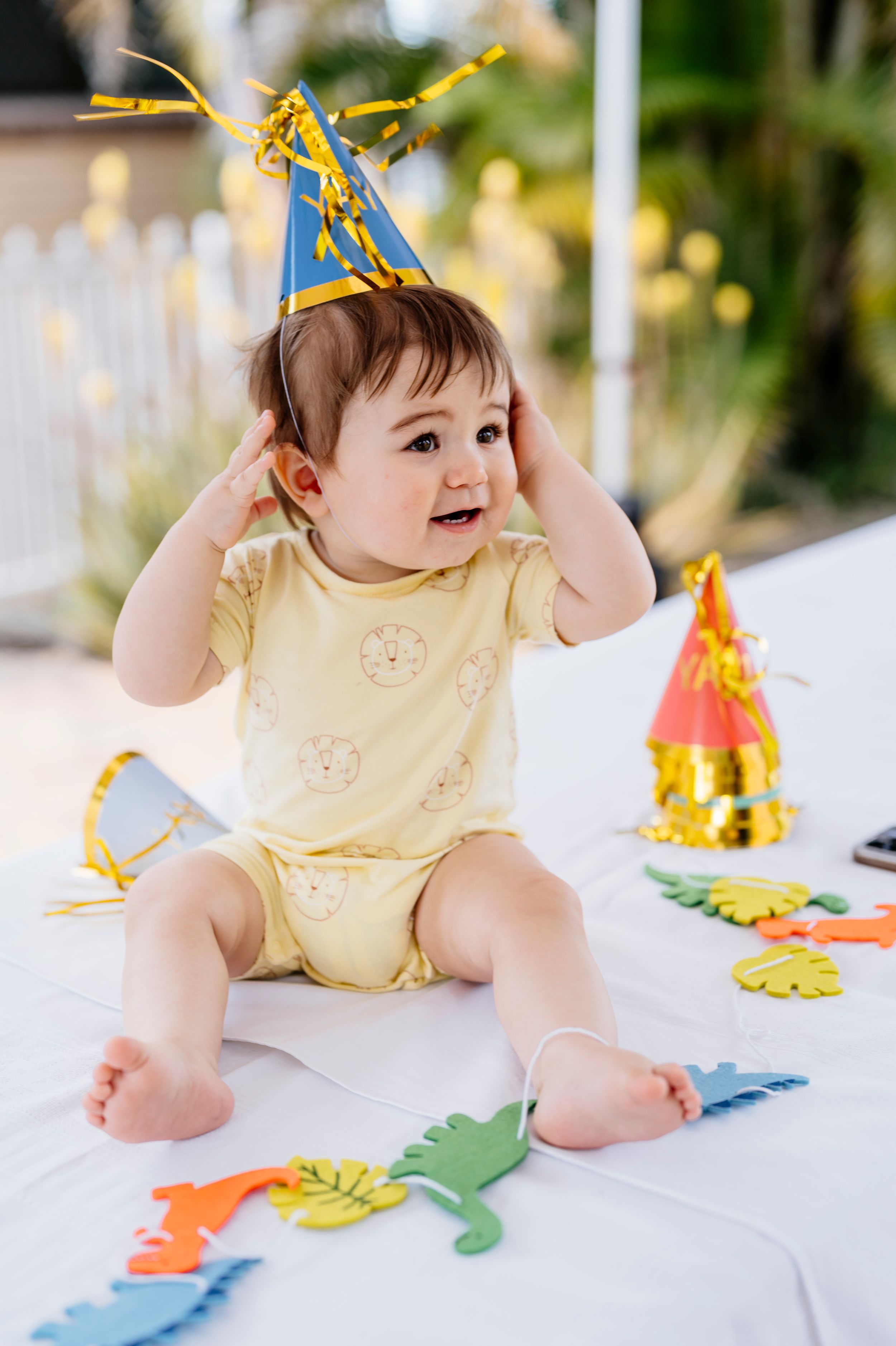 Baby wearing yellow romper and party hat sits on white surface with colorful party decorations.