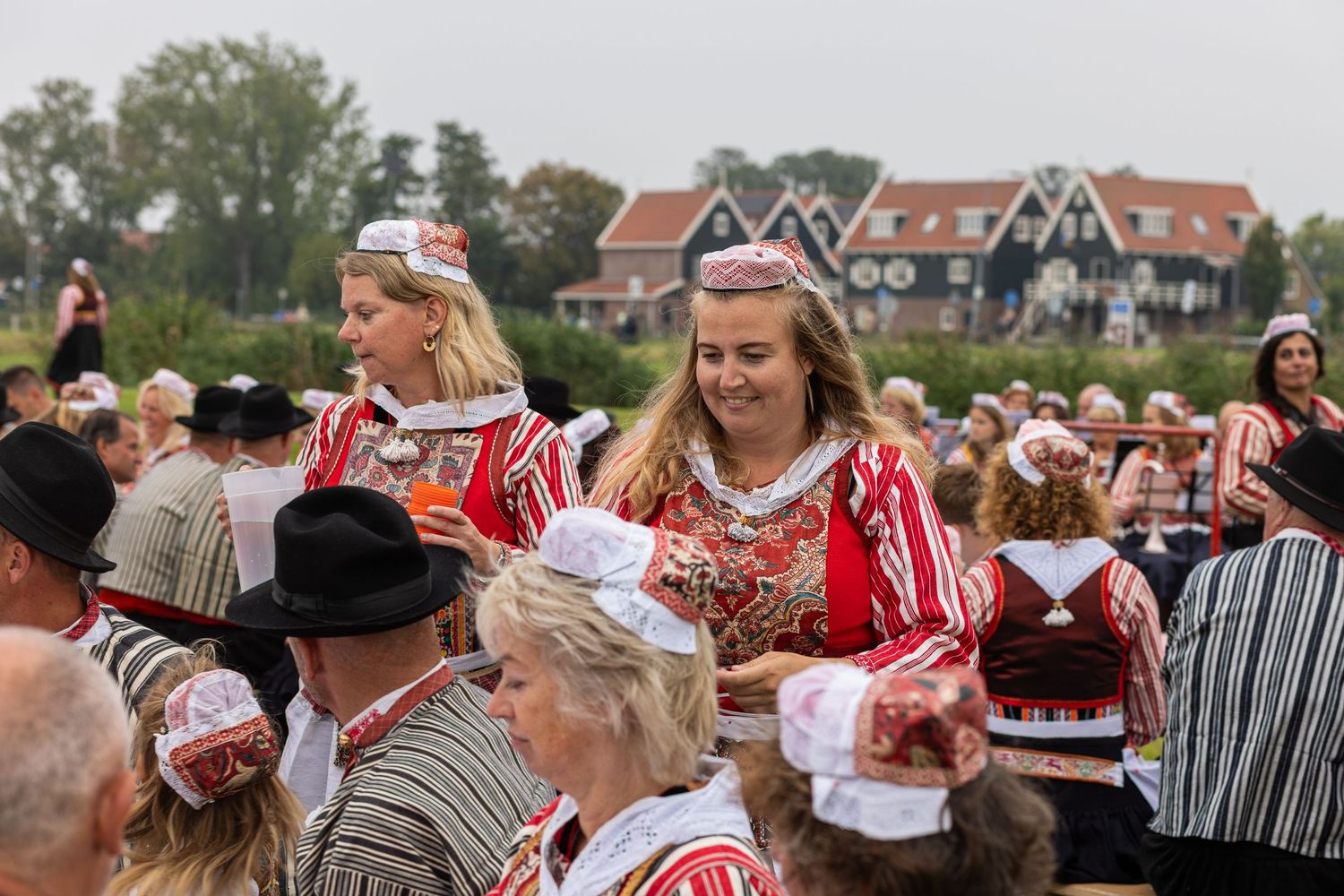 People in traditional Dutch costumes gather at an outdoor festival with trees and buildings in the background.