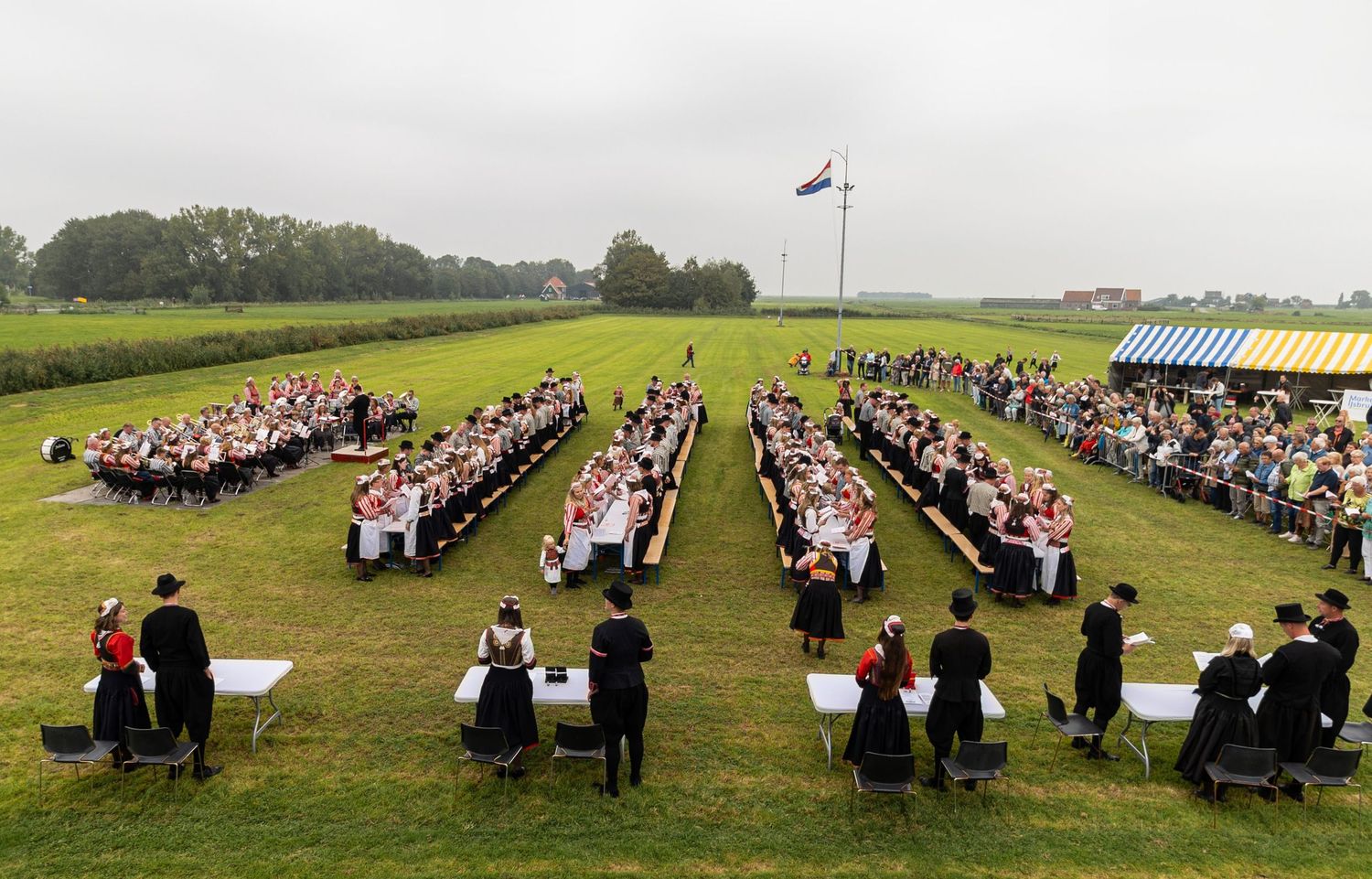 Large group of graduates in black caps and gowns sit in rows on a grassy field during an outdoor commencement ceremony.