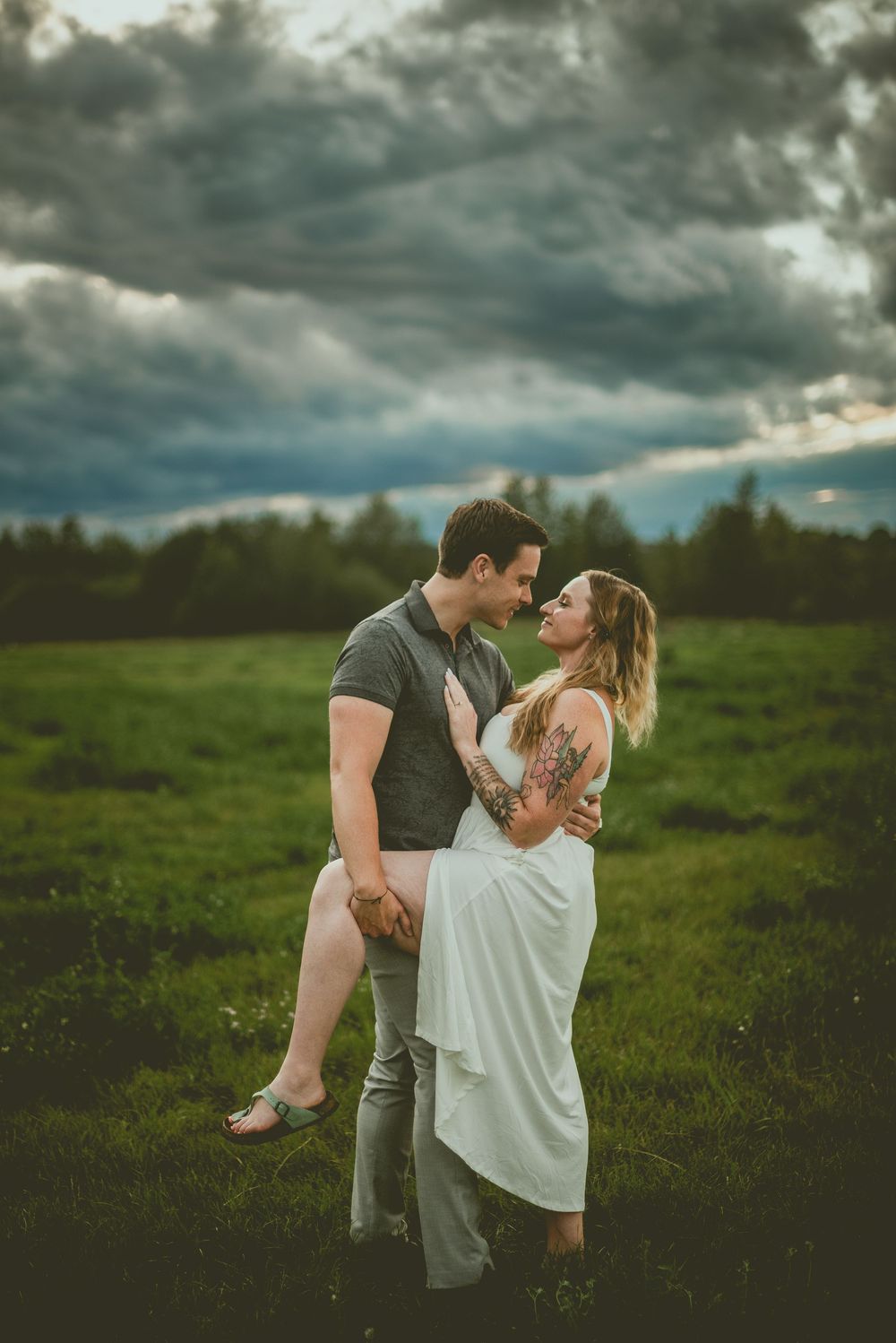 A couple embraces romantically in a lush green field under dramatic stormy clouds at dusk. Edmonton Couples Photography