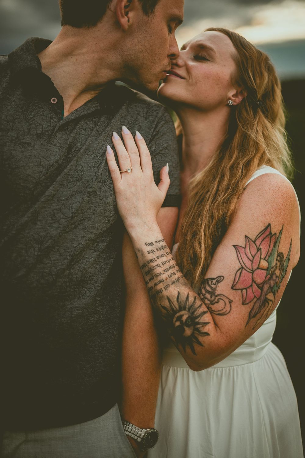 A couple in an intimate moment outdoors, showcasing a floral tattoo on her arm and text tattoo on his forearm. Edmonton Couples Photography