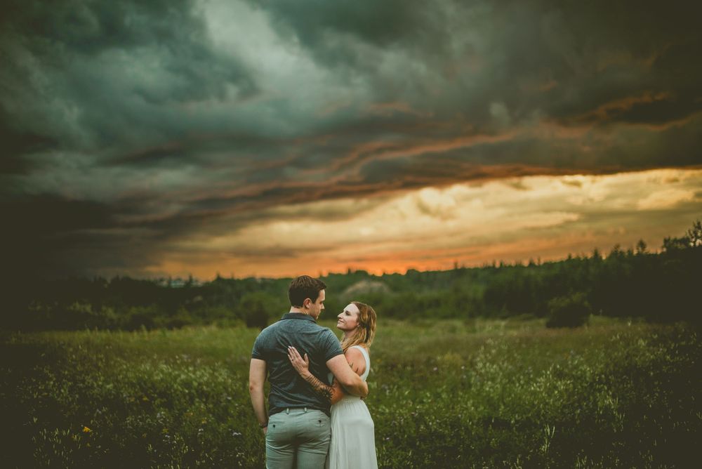 Couple embraces on hill during dramatic stormy sunset with dark clouds and golden sky over meadow. Edmonton Couples Photography