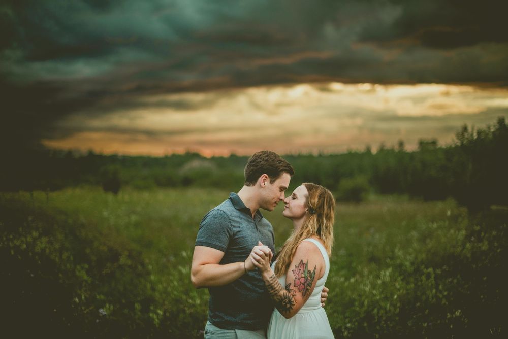 Romantic couple shares intimate moment in meadow at sunset while dark storm clouds gather overhead. Edmonton Couples Photography