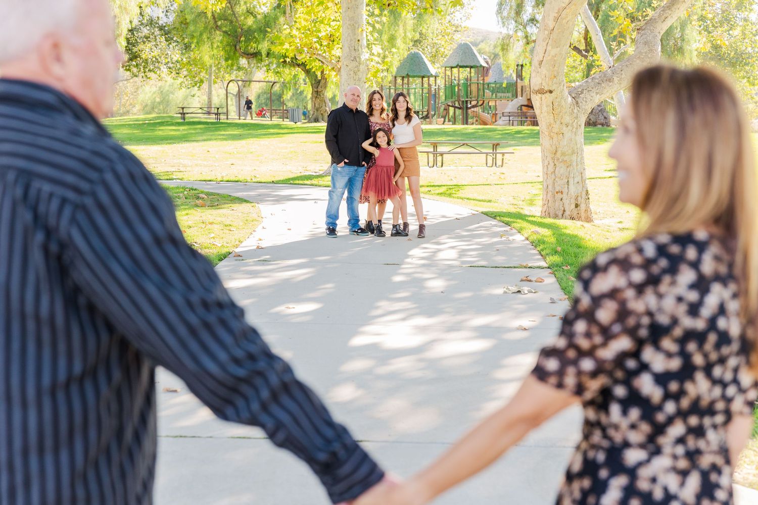 Two people hold hands and walk together on a sunny path through a park setting.