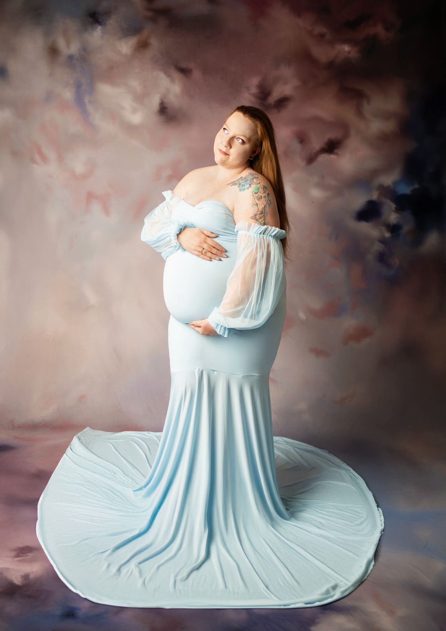 Maternity portrait featuring a woman in a flowing light blue gown against a dreamy painted backdrop.