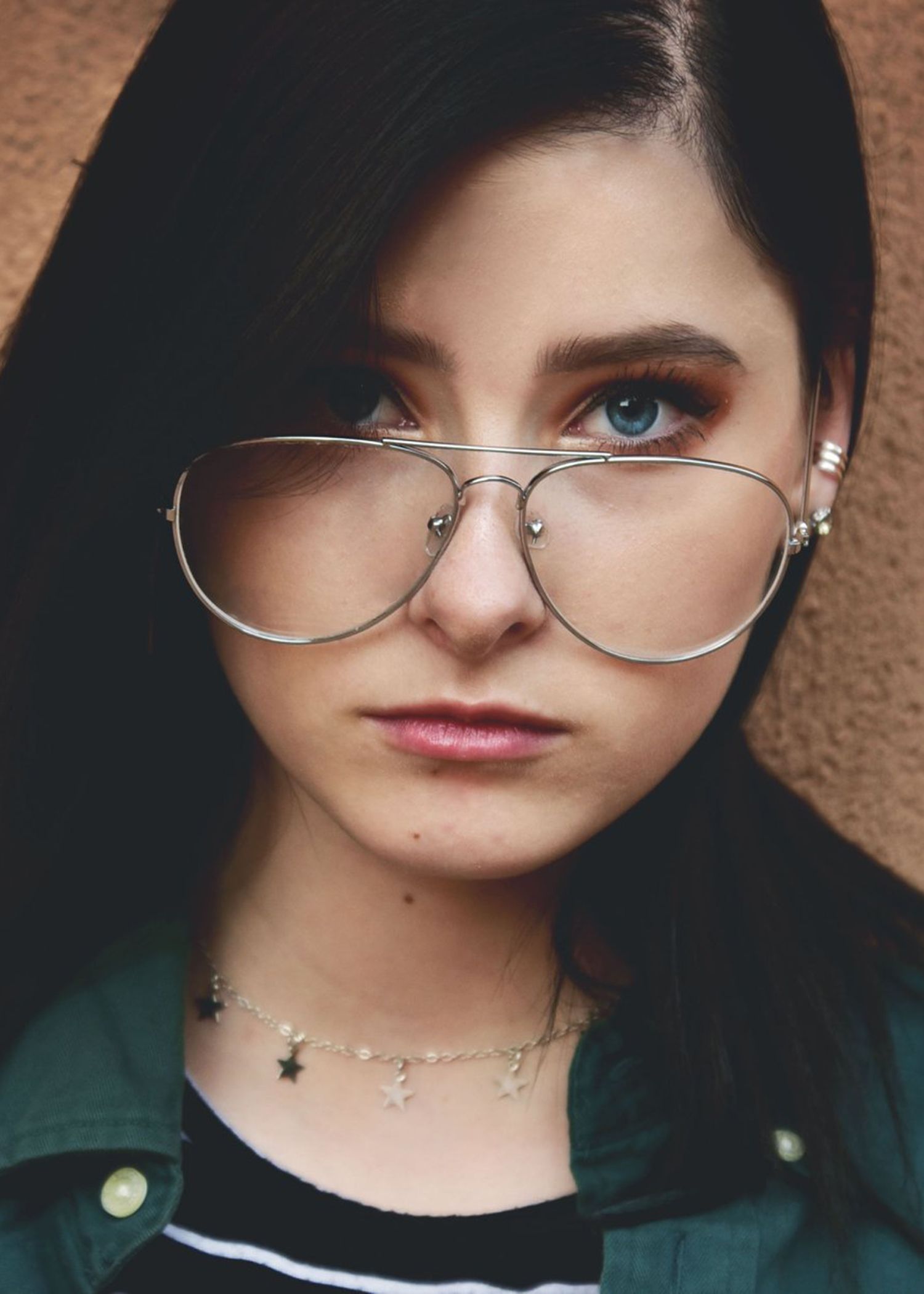 Portrait featuring aviator style glasses, delicate star necklace, and dark hair against textured brown background.