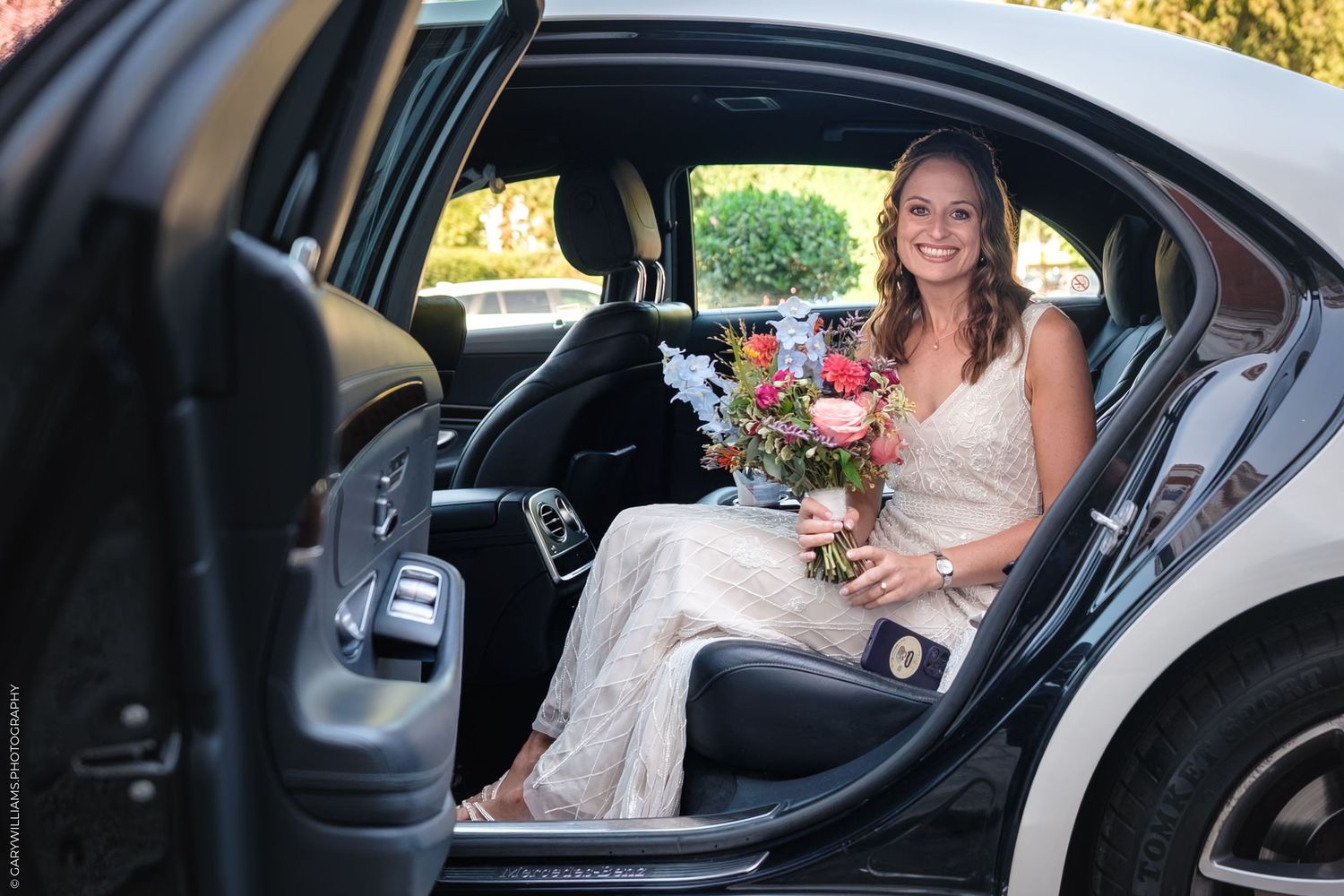 A person in formal dress texting while seated in a car's passenger seat on a sunny day.