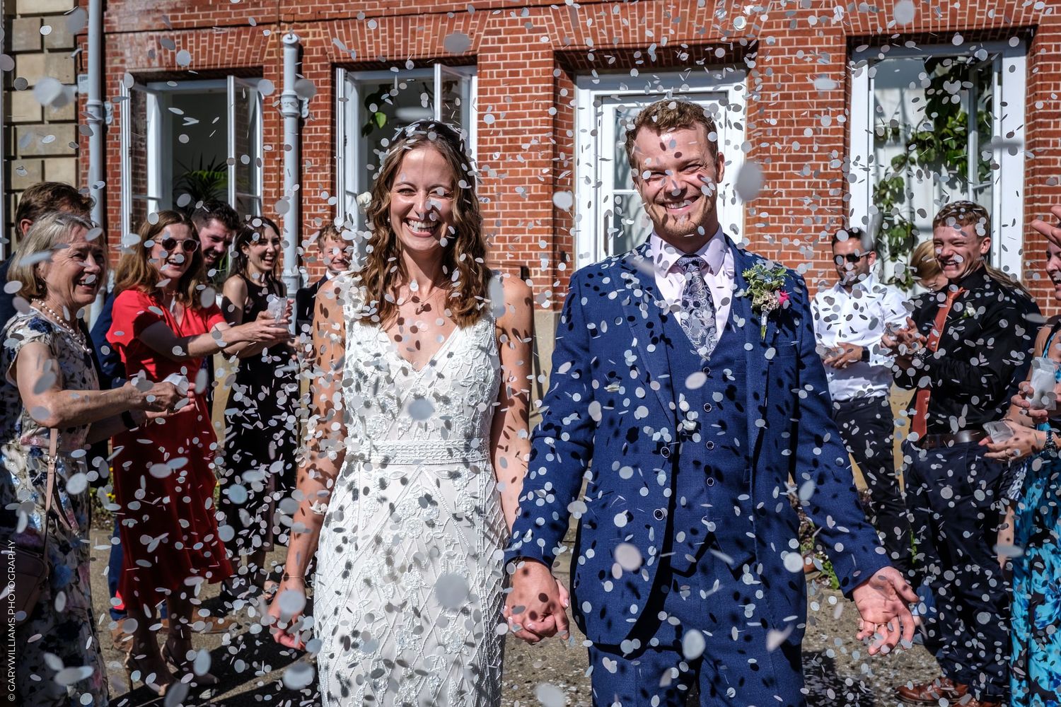 Newlyweds exit church through cloud of white confetti as guests celebrate on brick building steps.