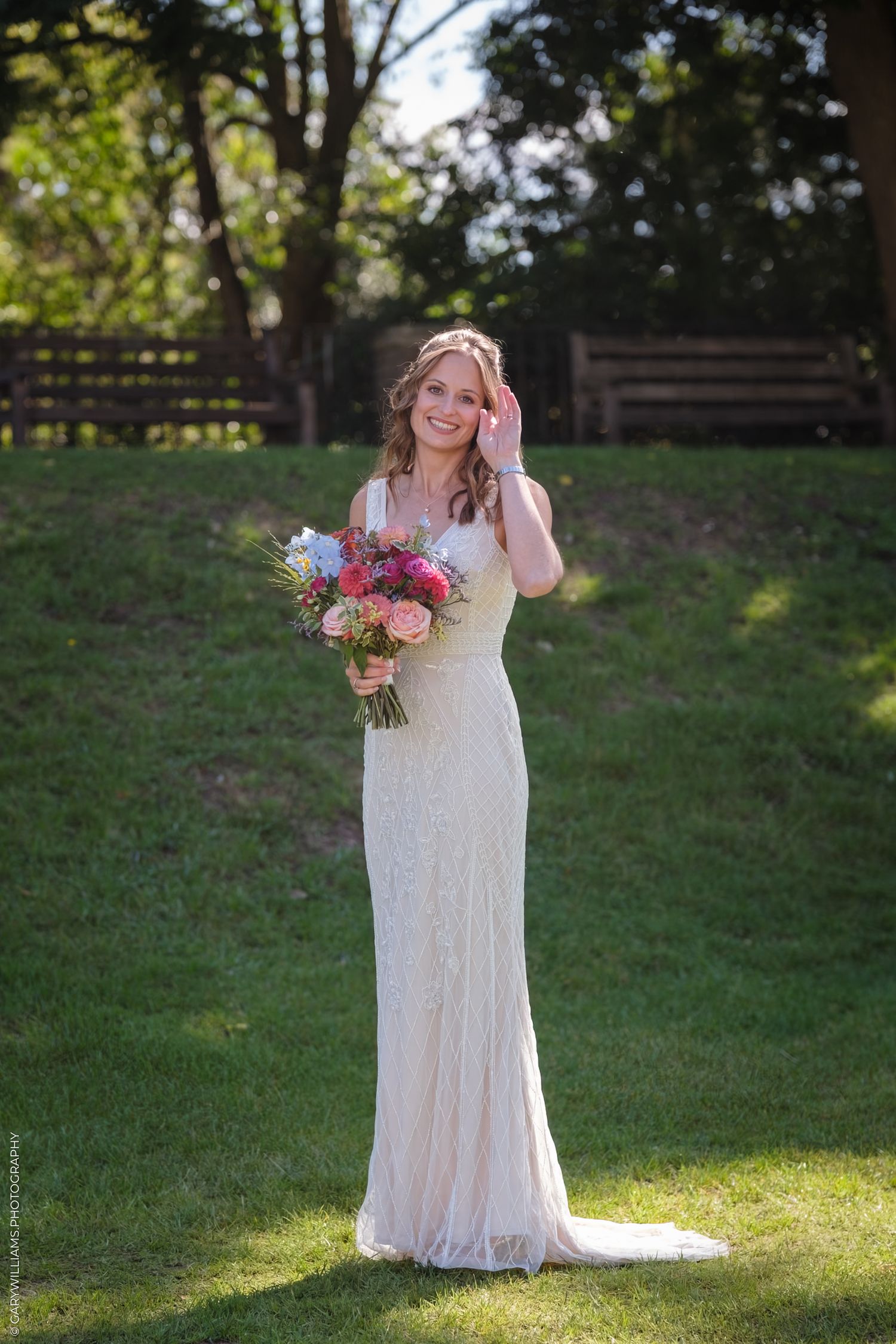 Bride in a lace white wedding dress carries a colorful bouquet while posing outdoors in a garden setting.