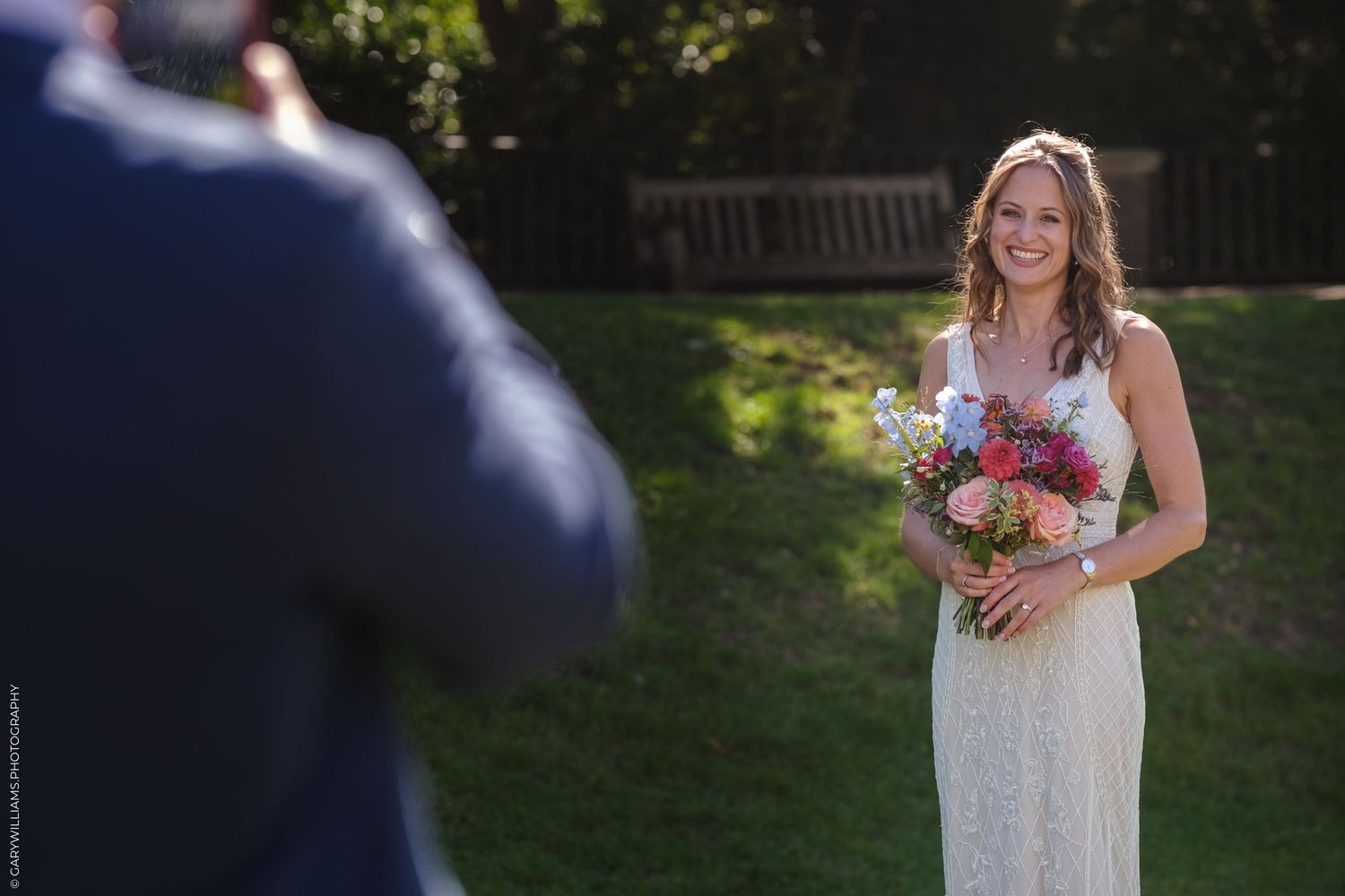 A bride in a white lace wedding dress holds a vibrant bouquet in a sun-dappled garden setting.