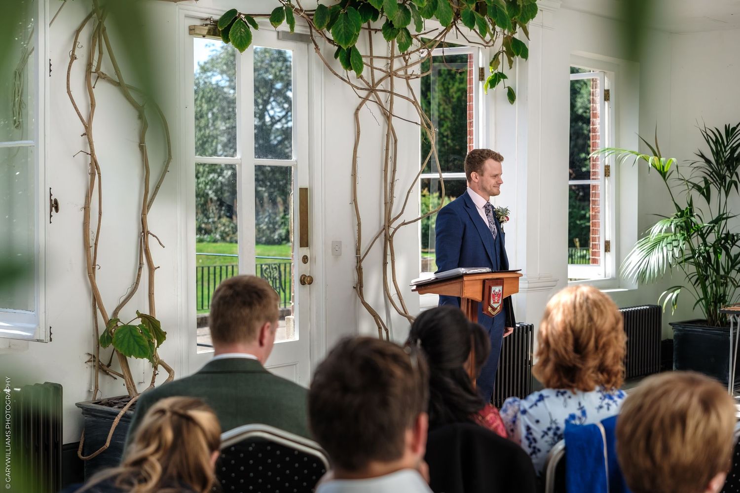 Wedding ceremony guests sit in white chairs surrounded by green plants and vines in a bright, airy indoor venue.