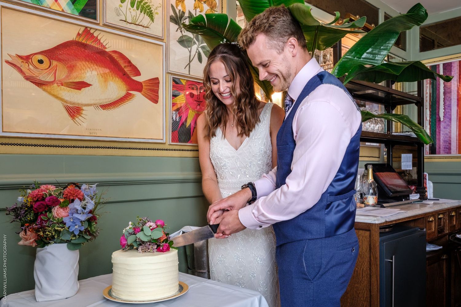 A couple cuts into their wedding cake together in a cozy restaurant setting with a large fish painting on the wall.