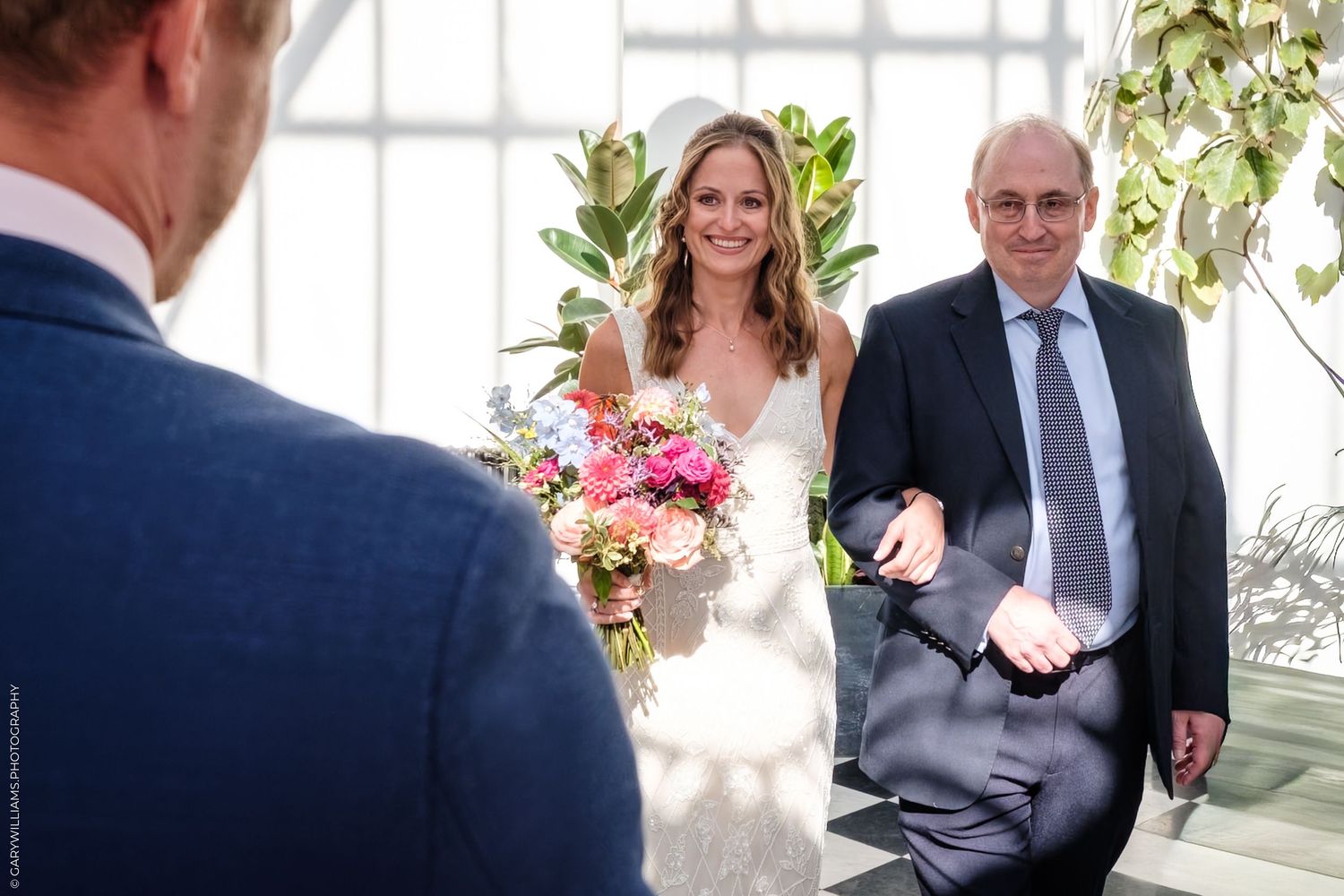 A bride in a white wedding dress holding pink bouquet walks down the aisle with escort in a greenhouse setting.