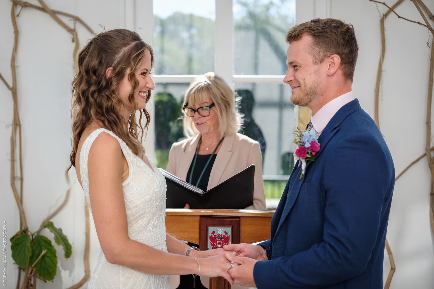 A couple holds hands during a ceremony while standing in front of an officiant inside an elegant white venue.