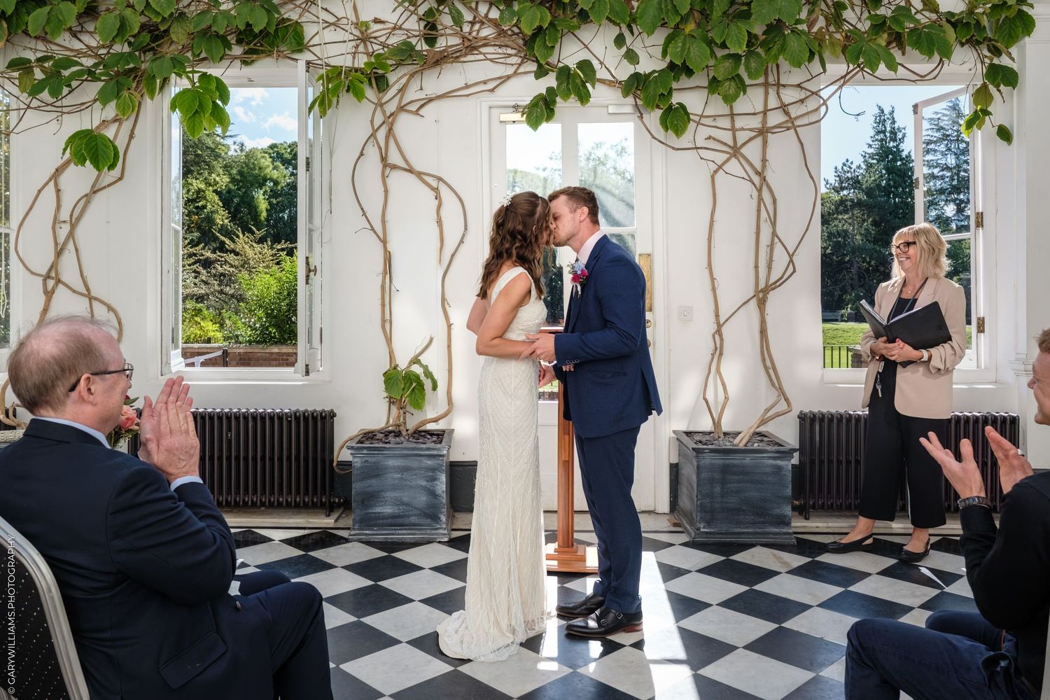 Wedding ceremony taking place in a gorgeous indoor venue with black and white checkered floors and ivy-covered columns.