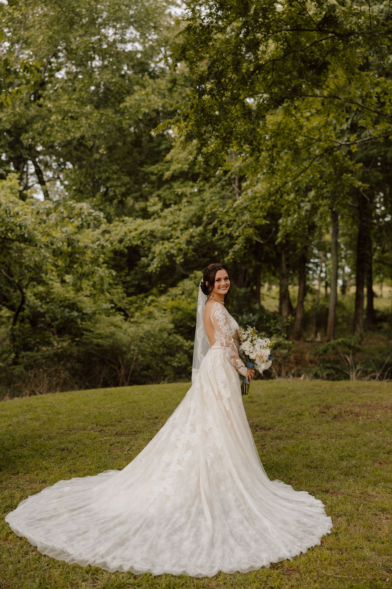 Bride in lace wedding gown with long train and veil holds bouquet, smiling over shoulder in lush green garden.