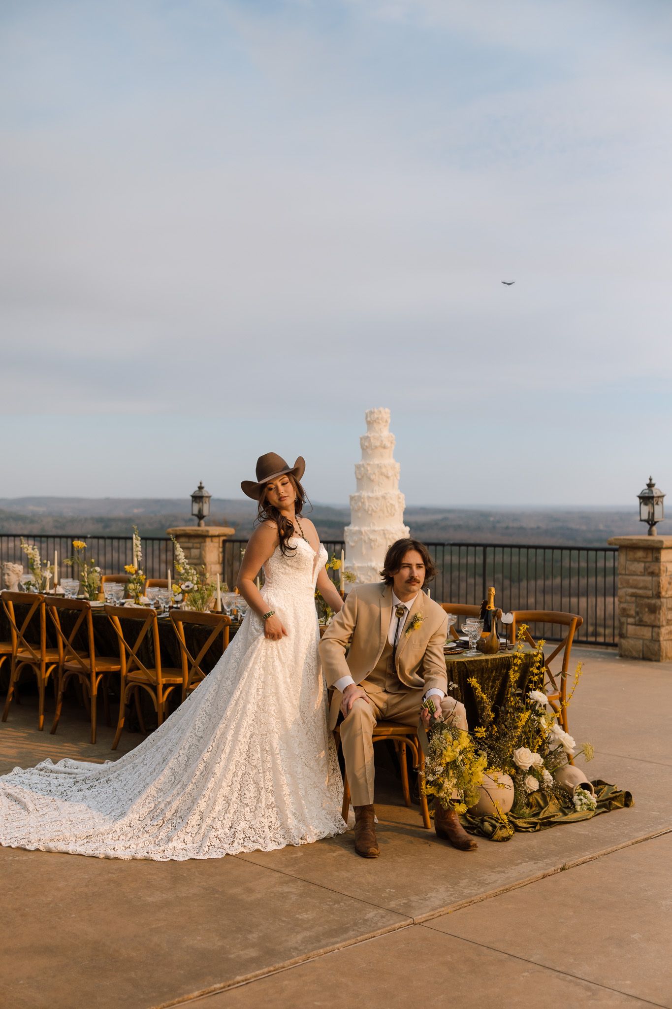 Bride in lace gown and cowboy hat poses with groom in tan suit at outdoor wedding reception with tiered cake.