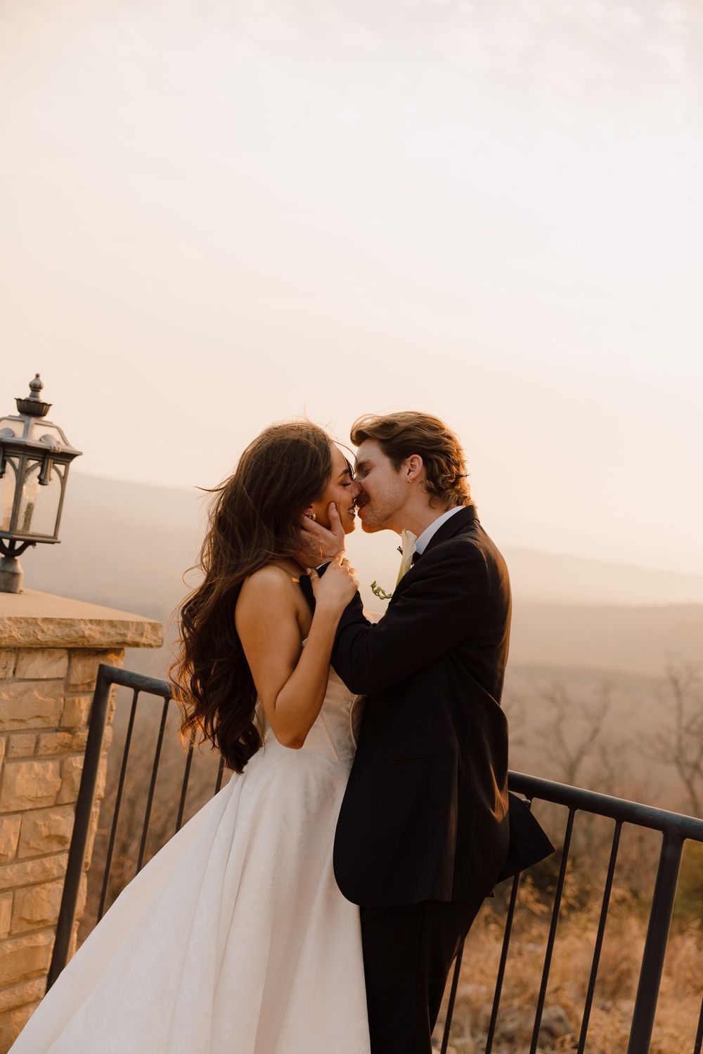 A bride and groom share a romantic kiss on a balcony at sunset with scenic mountain views behind them.