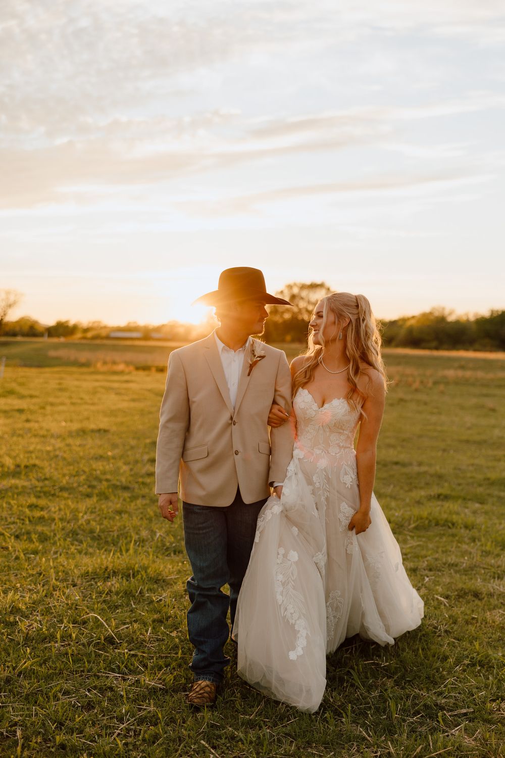 Bride and groom walking at sunset in a green field, she in lace gown, he in tan blazer and cowboy hat.