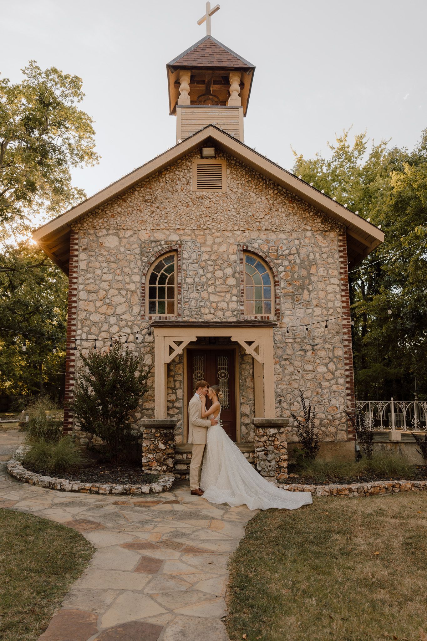 A rustic stone chapel with arched windows and bell tower serves as a romantic wedding venue in a wooded setting.