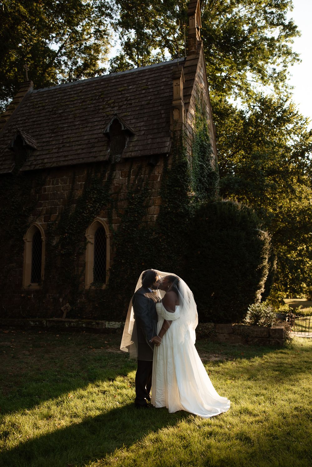 Newlywed couple sharing a romantic kiss in front of a charming ivy-covered stone chapel surrounded by lush greenery.