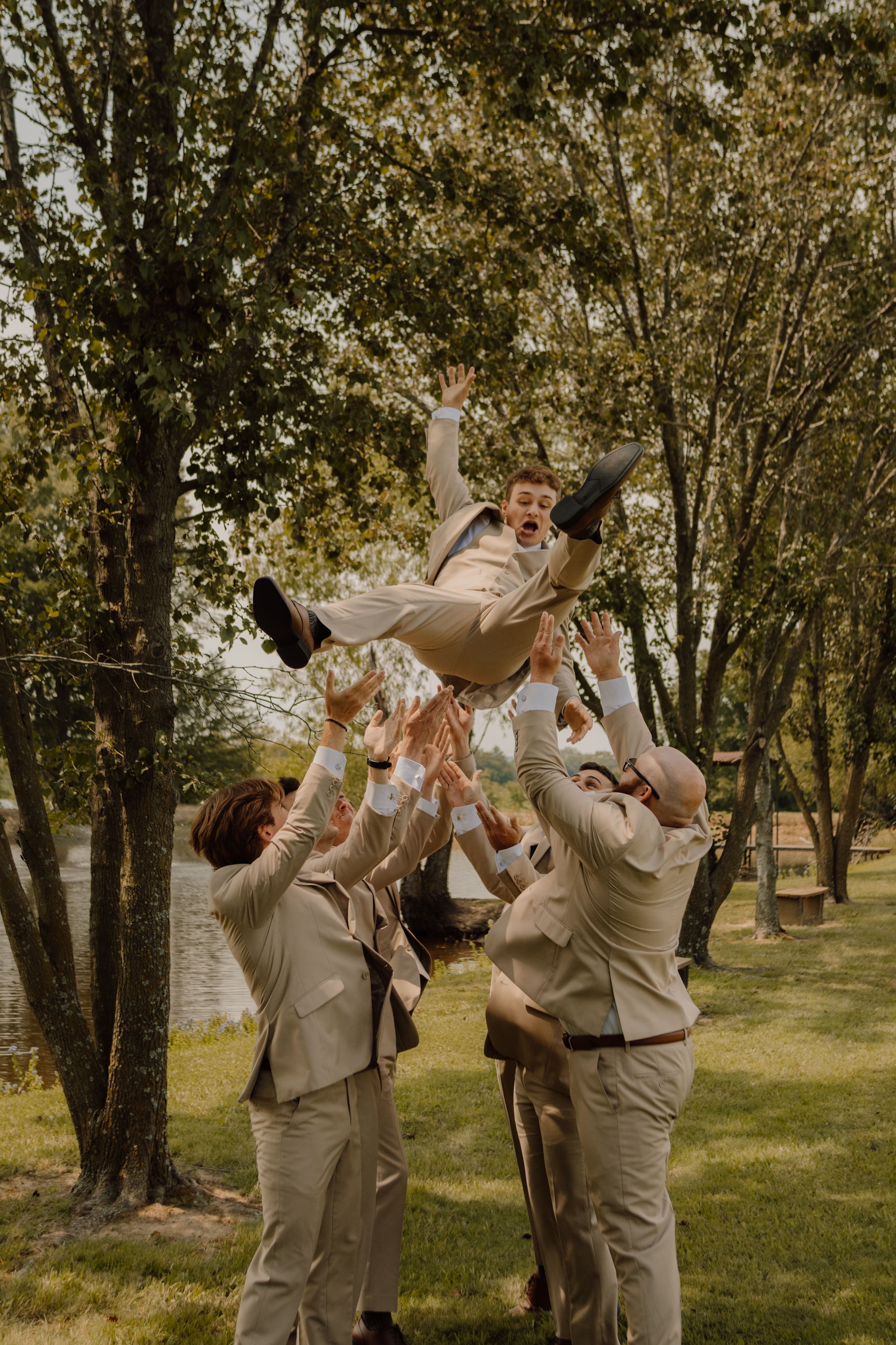 A group in beige suits celebrate and toast together with drinks in a wooded outdoor setting during an event.