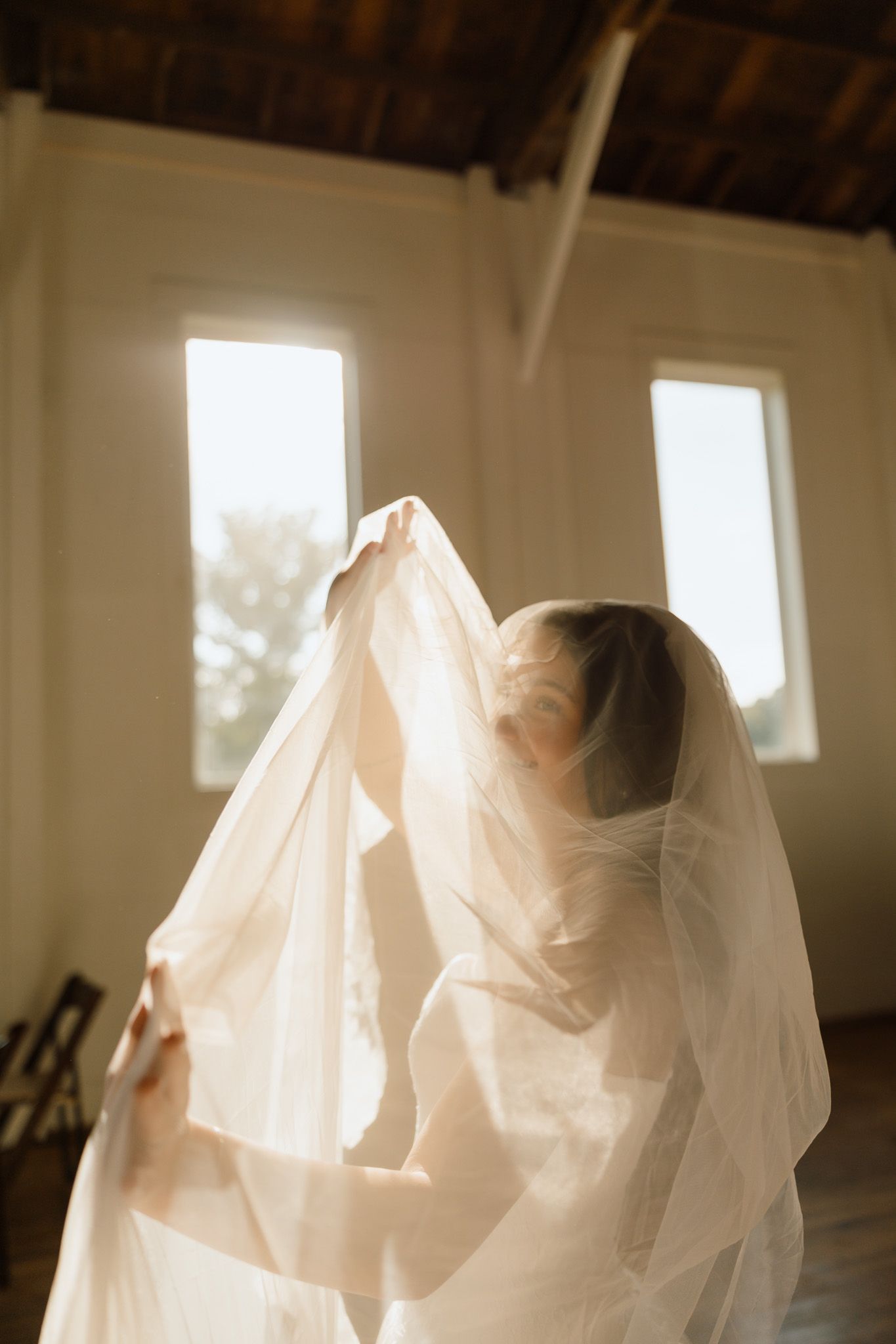 Bride lifting her sheer wedding veil in golden backlit light inside a rustic venue with wooden ceiling.