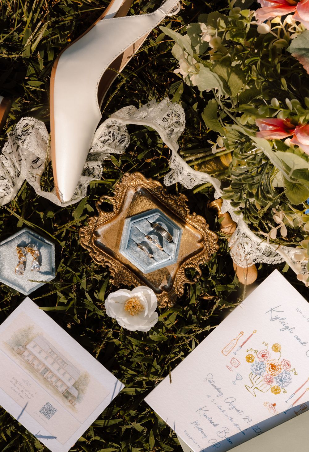 Wedding flat lay with gold ring dish, rings, bridal heels, lace, florals, and illustrated stationery on green grass.