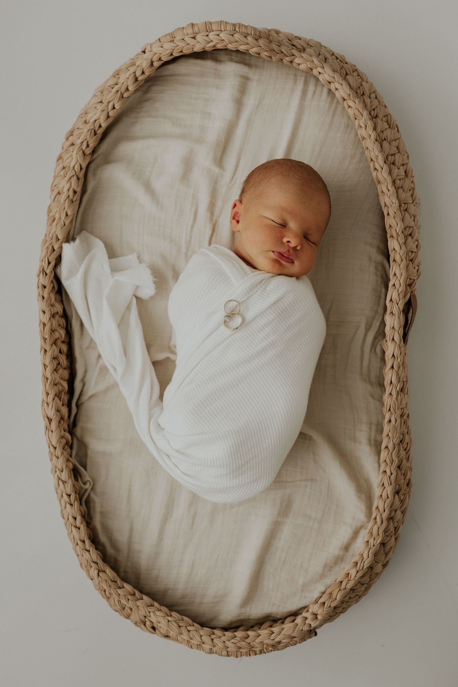 A sleeping newborn baby swaddled in white lies peacefully in an oval woven basket with neutral bedding.