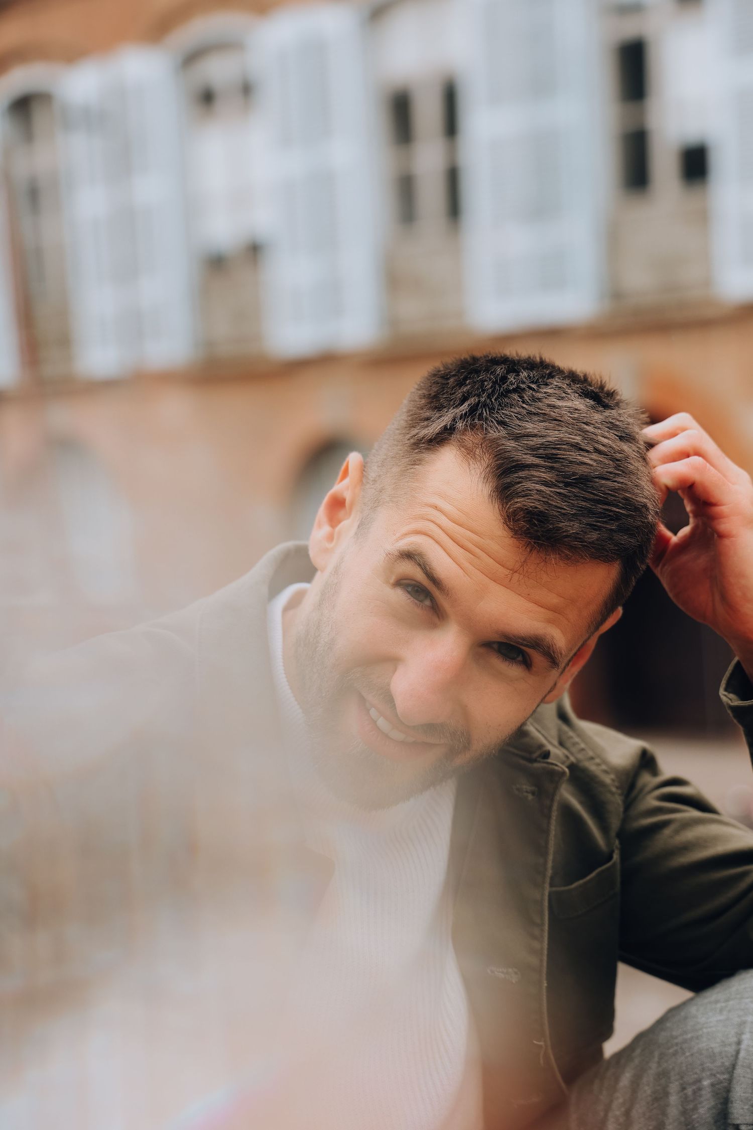 A smiling man in a green jacket runs his hand through his hair outdoors near a historic building.