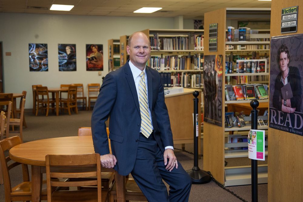 A person in a navy suit and striped tie stands in a school library with bookshelves and tables visible.
