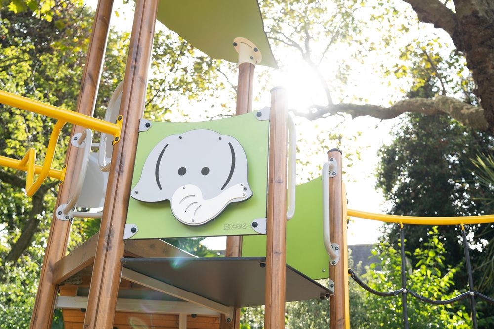 A playground structure with a cute elephant face decoration panel on a green background, illuminated by sunlight through trees.