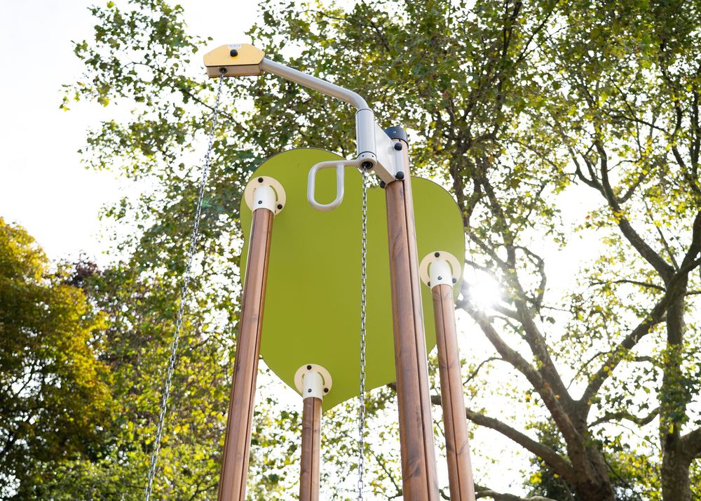 Green and brown playground equipment with spiral climbing elements stands tall against a backdrop of leafy trees in a park setting.