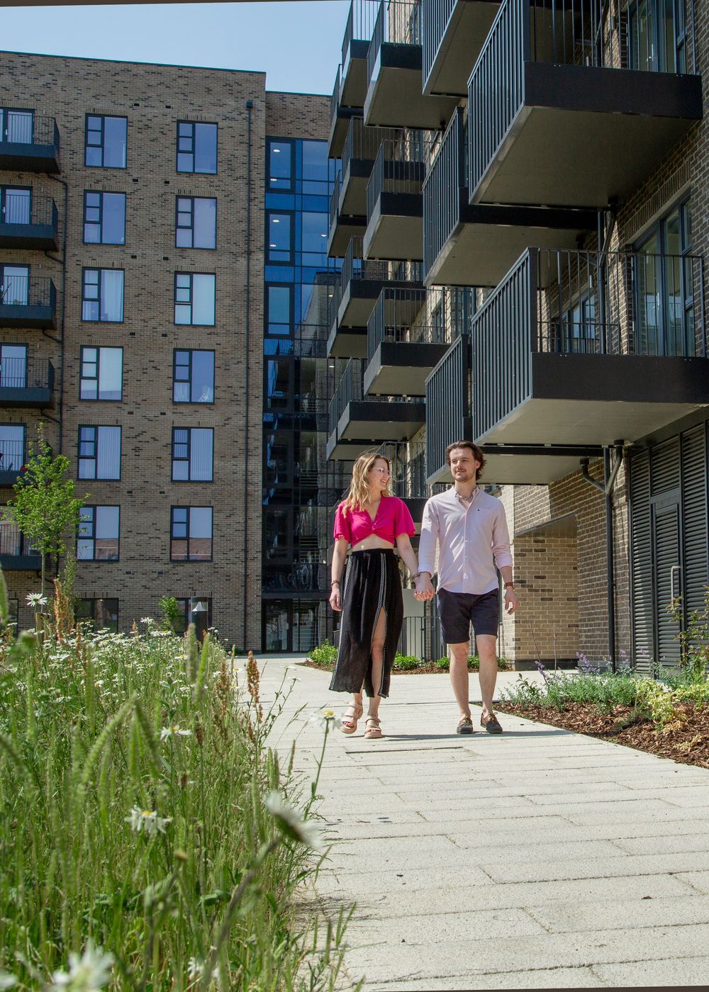 A couple walks on a concrete pathway alongside a modern brick apartment building with balconies and landscaped greenery.