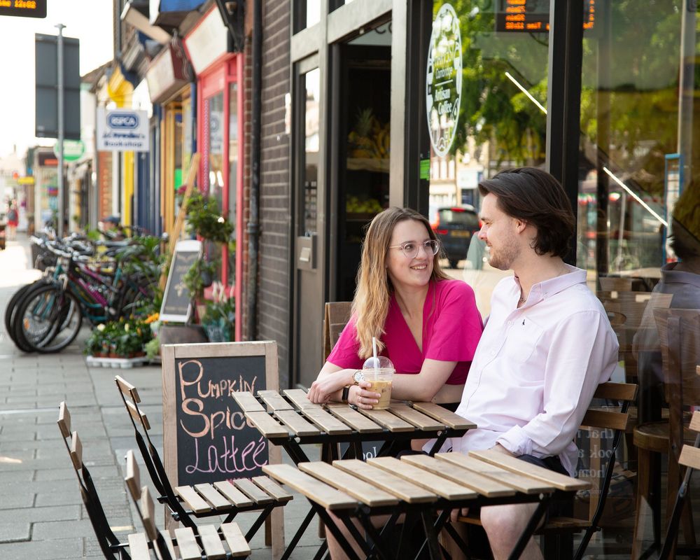 A group of people sit and chat at outdoor cafe tables along a city sidewalk on a sunny day.