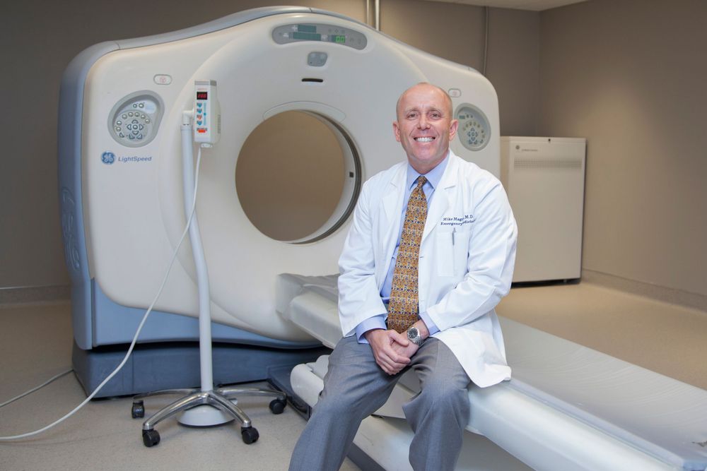 Medical professional sitting beside CT scanning machine in clinical setting.