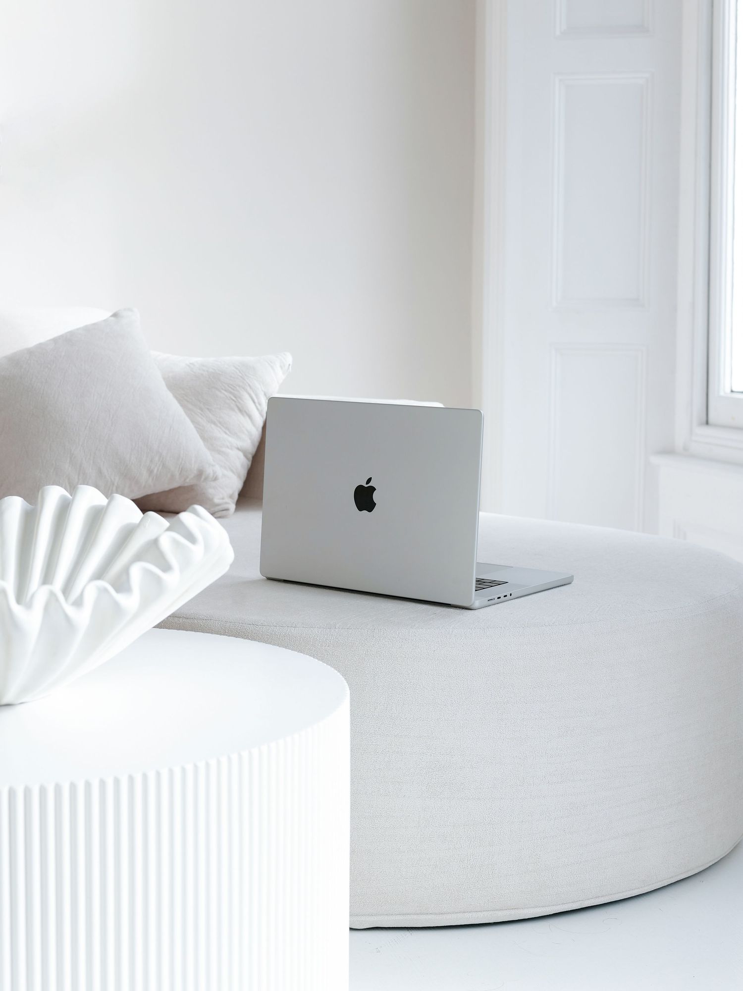 Silver MacBook laptop on a round white ottoman in a minimalist all-white living room with cushions and natural light.