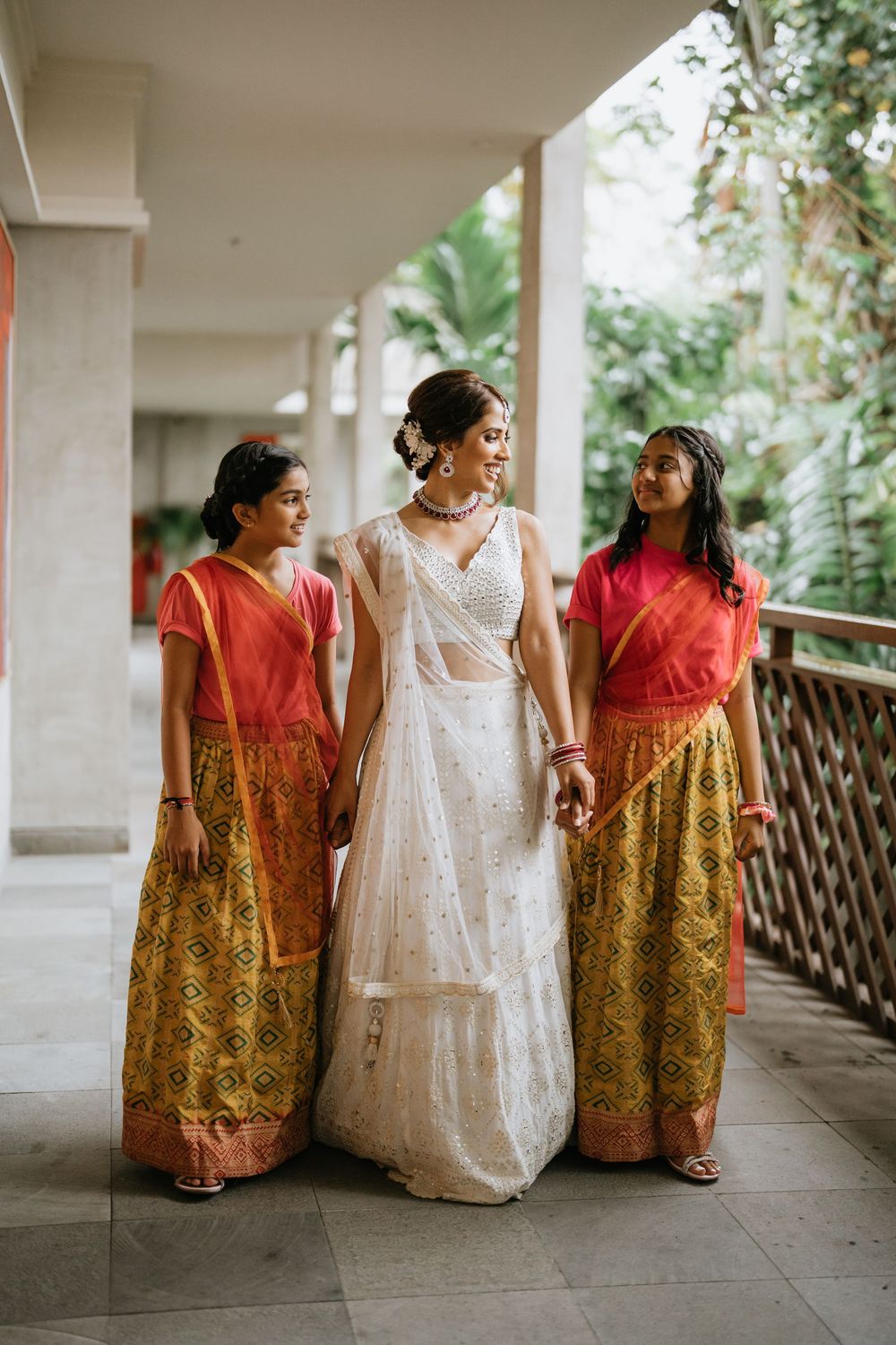 A group wearing traditional Indian wedding attire with red and gold saris and a white dress pose together in a covered walkway.