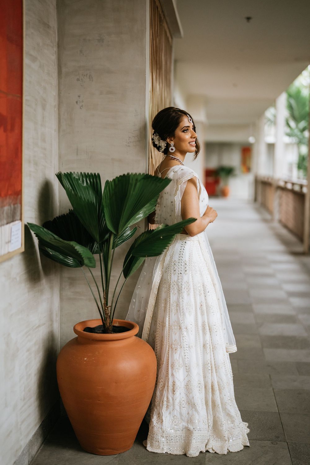 A white wedding dress is showcased against a backdrop of potted palm plants in an elegant covered corridor.